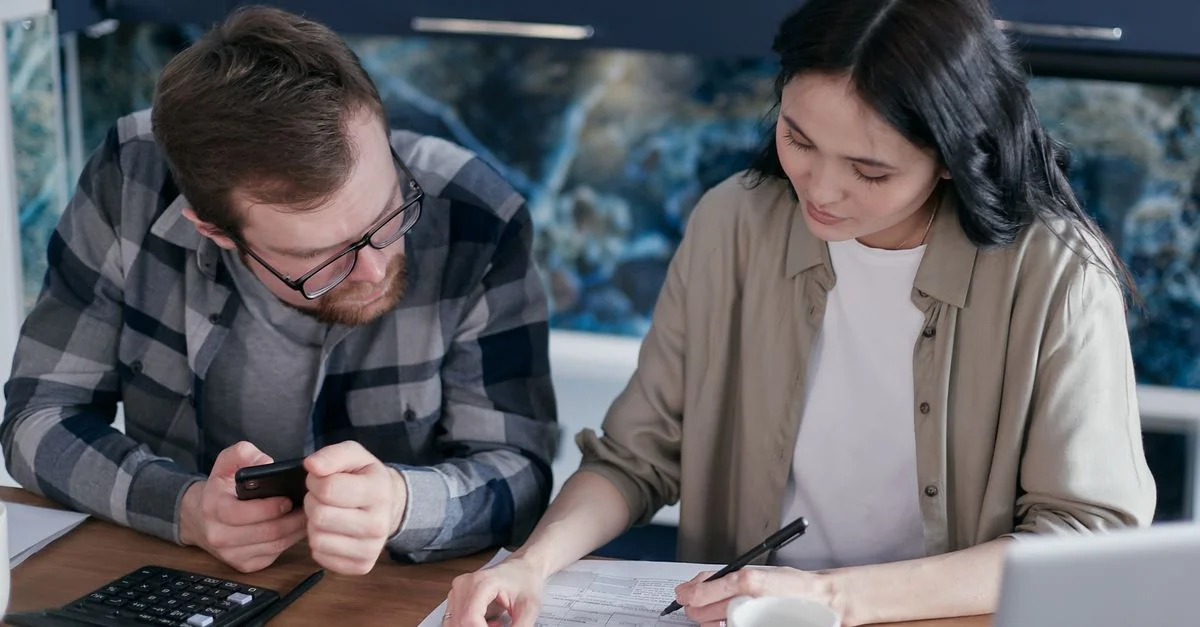 Un couple examinant des documents financiers à une table, utilisant des gadgets comme une calculatrice et un téléphone.