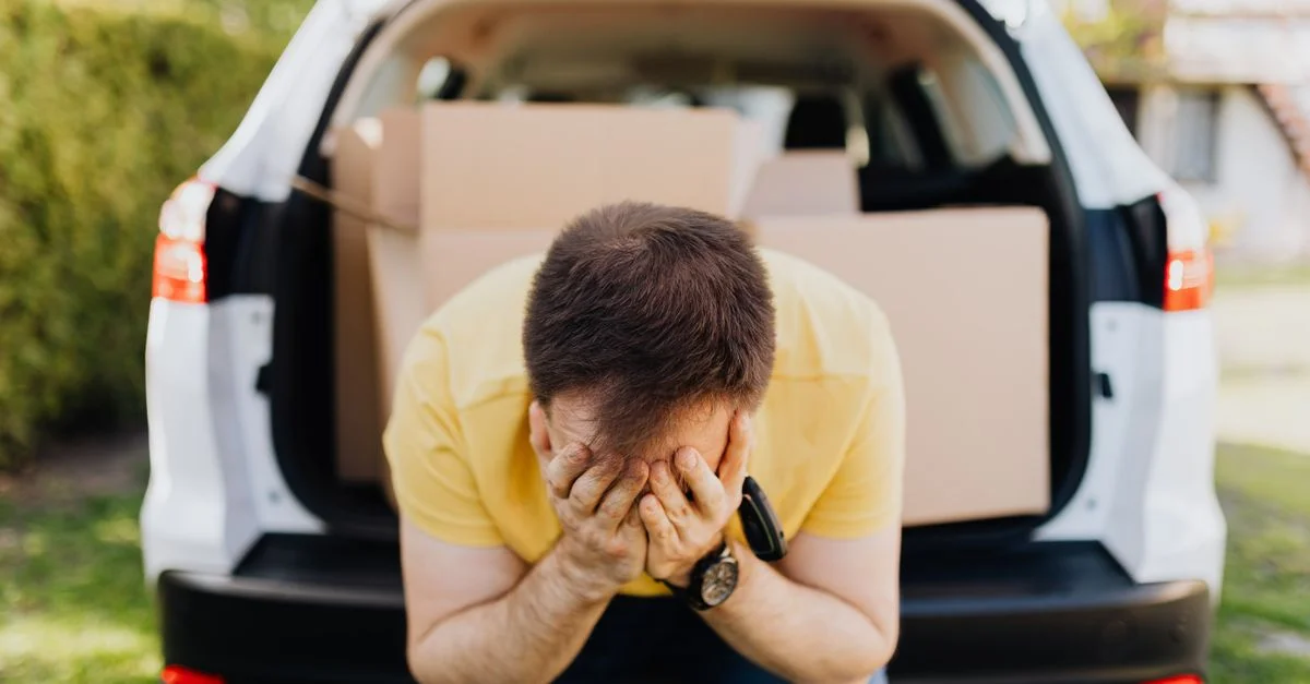 Un homme en détresse assis à côté d'un coffre de voiture rempli de cartons, exprimant du stress.