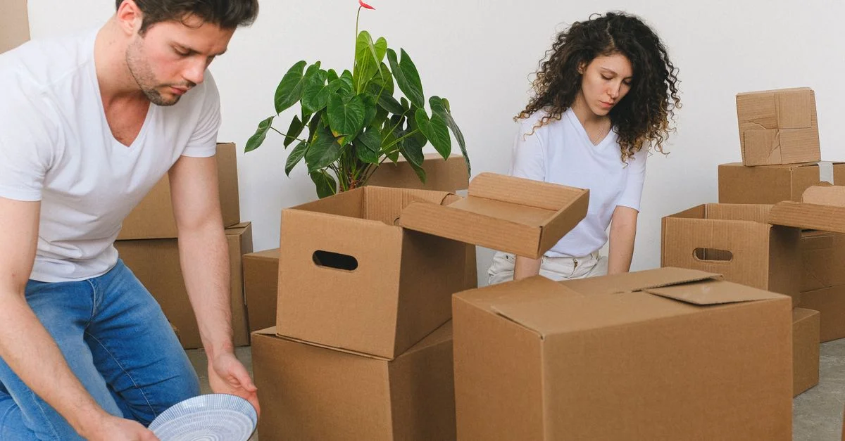Un jeune couple range des cartons en déménageant dans un nouvel appartement. Des cartons en carton et une plante sont visibles.